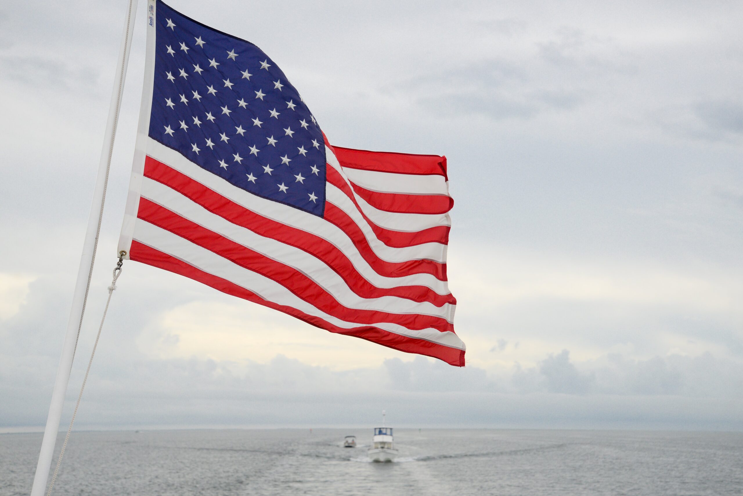 A vibrant American flag flutters on a boat with a vast ocean and cloudy sky backdrop.