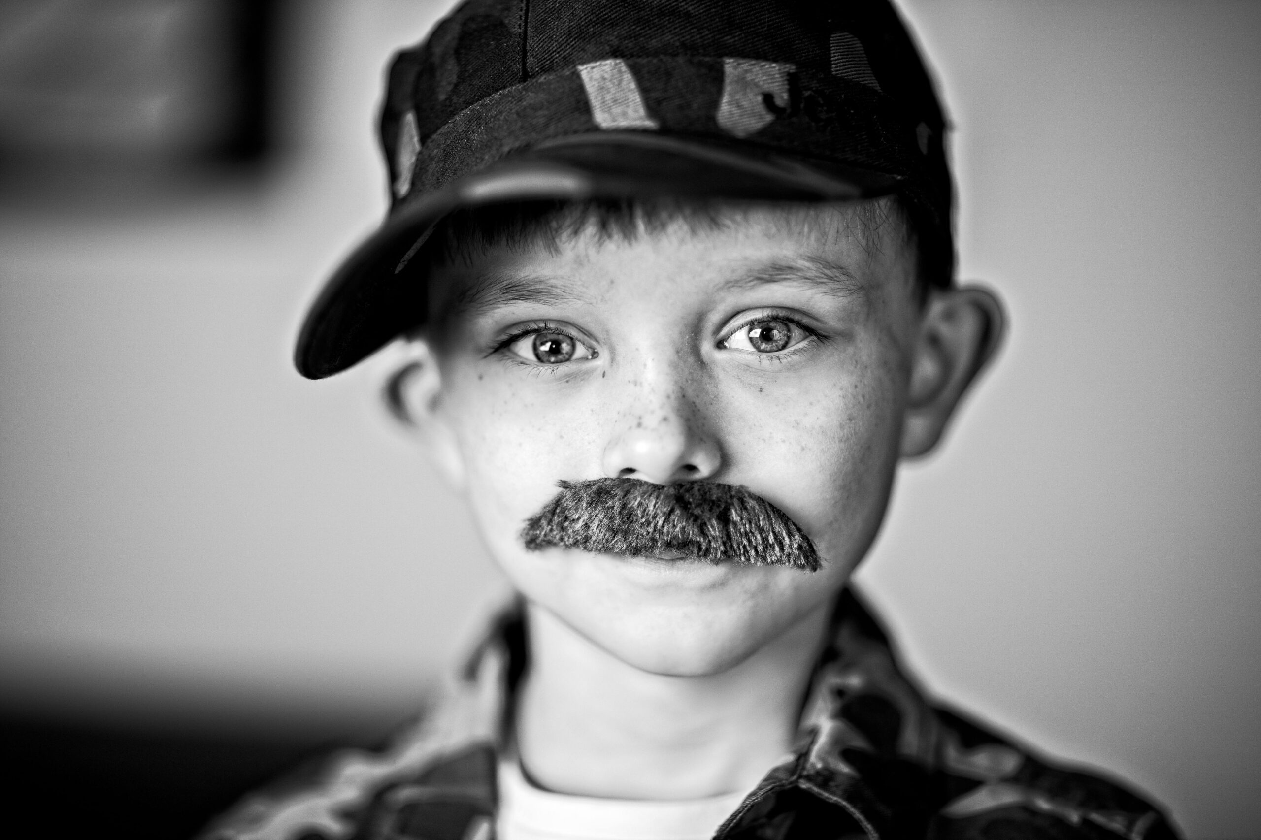 A grayscale portrait of a young boy wearing a cap and a fake mustache. Captures the playful essence of youth.