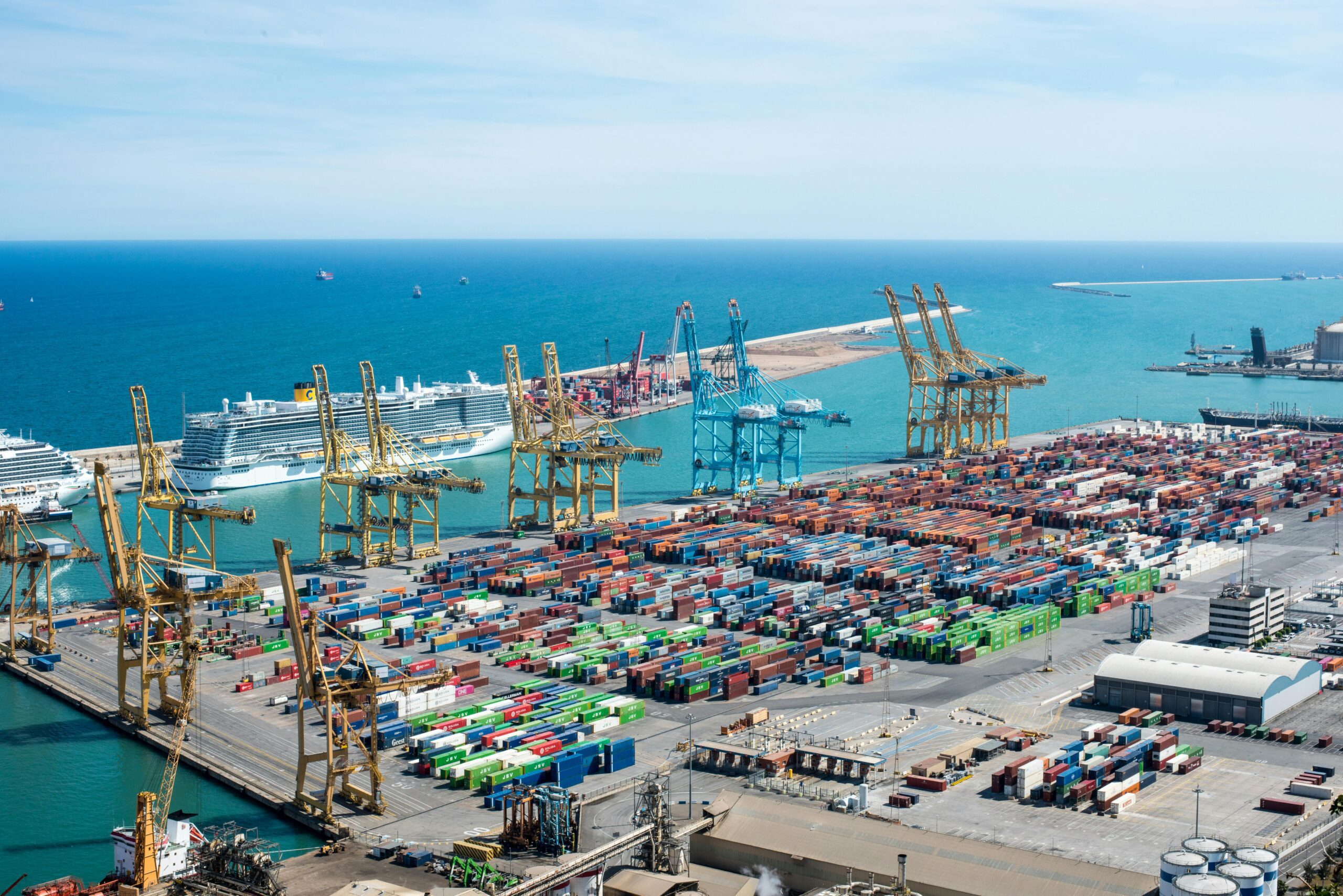A vibrant aerial shot of Barcelona port featuring cranes, containers, and cruise ships on a sunny day.