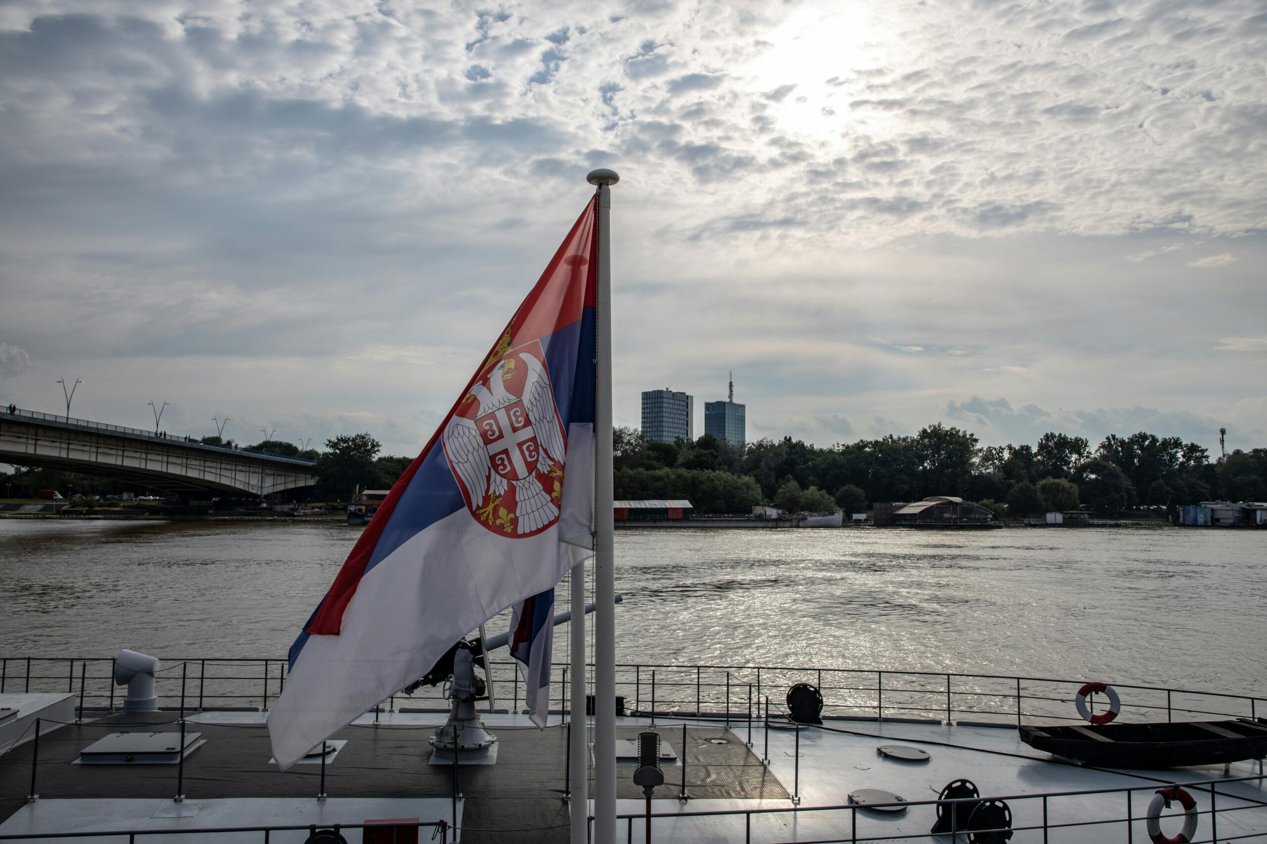 Serbian flag on a boat with an urban skyline and river scene in daytime.