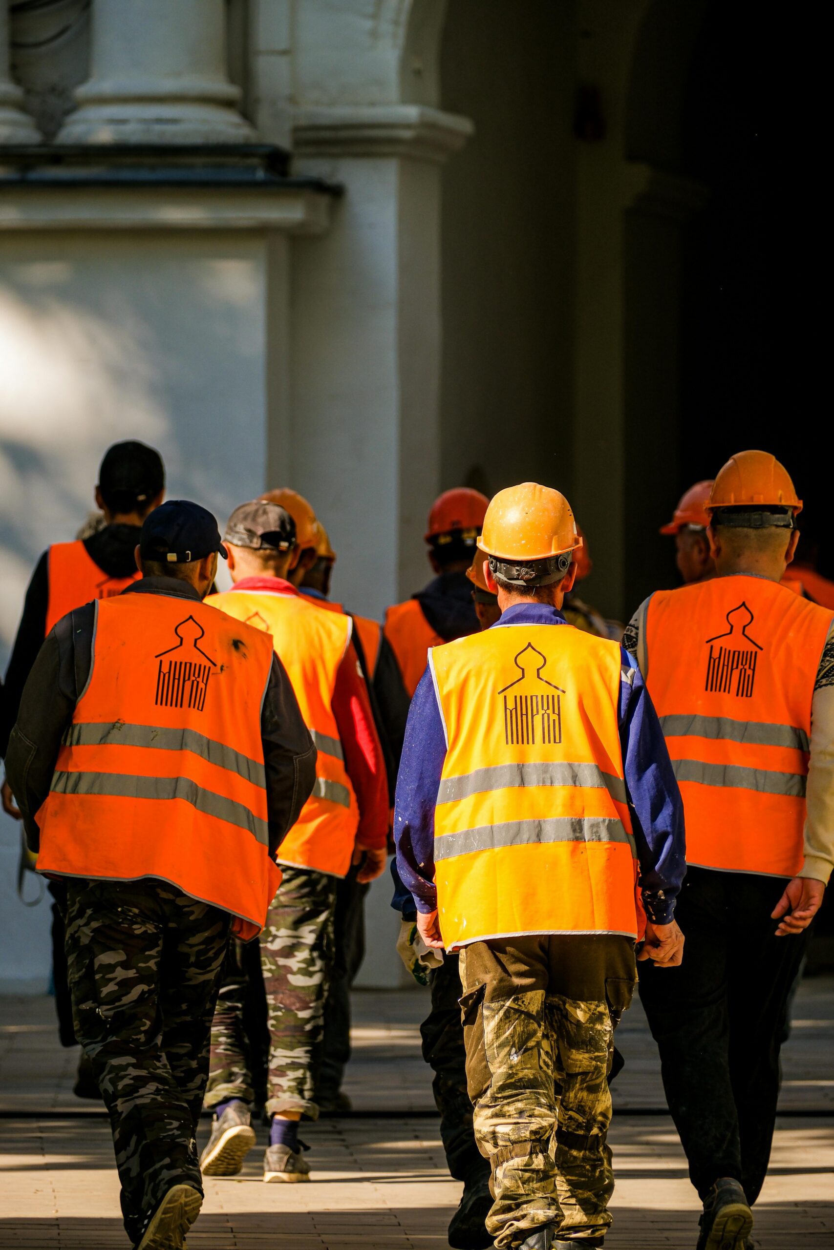 Group of construction workers wearing orange vests and helmets walking in urban area.