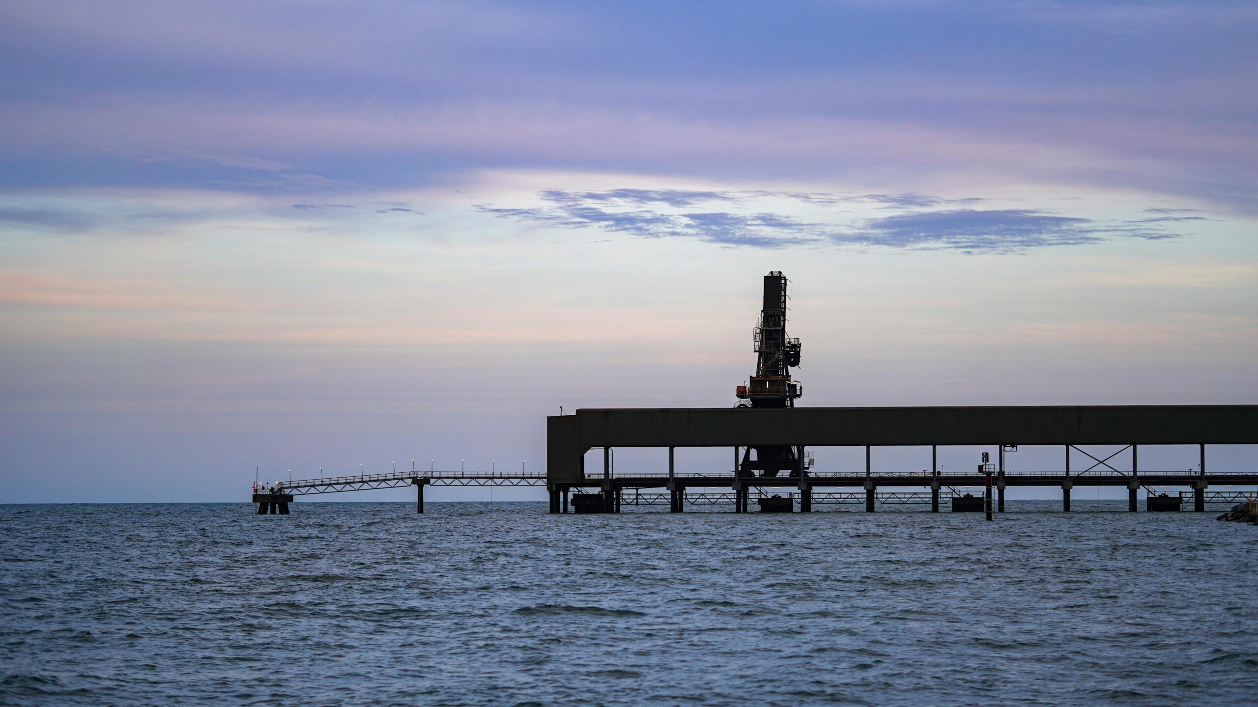 An industrial pier extends over a calm sea with a beautiful sunset sky.