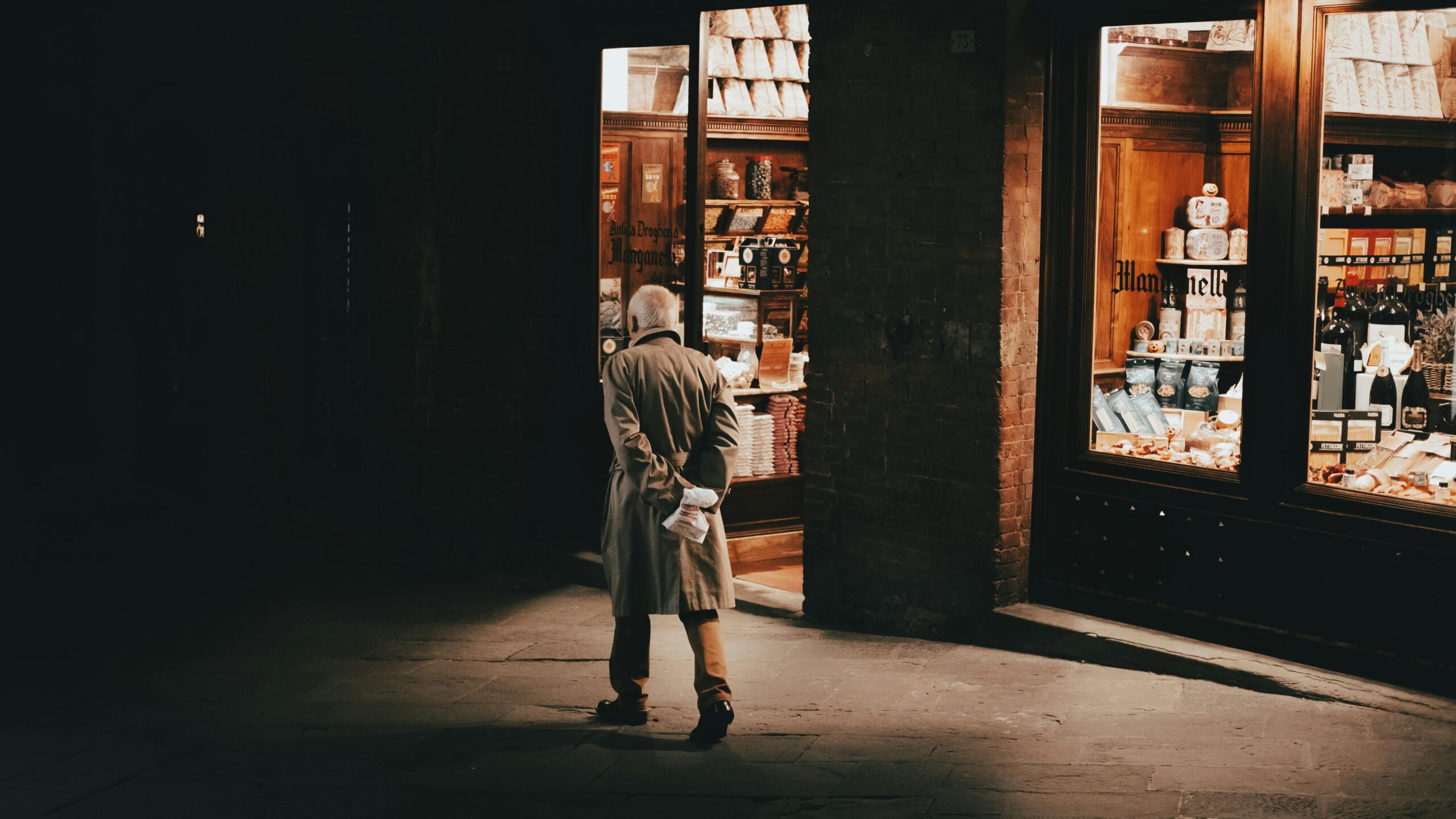A senior man in a trench coat looks into a warmly lit shop window at night, creating a moody street scene.