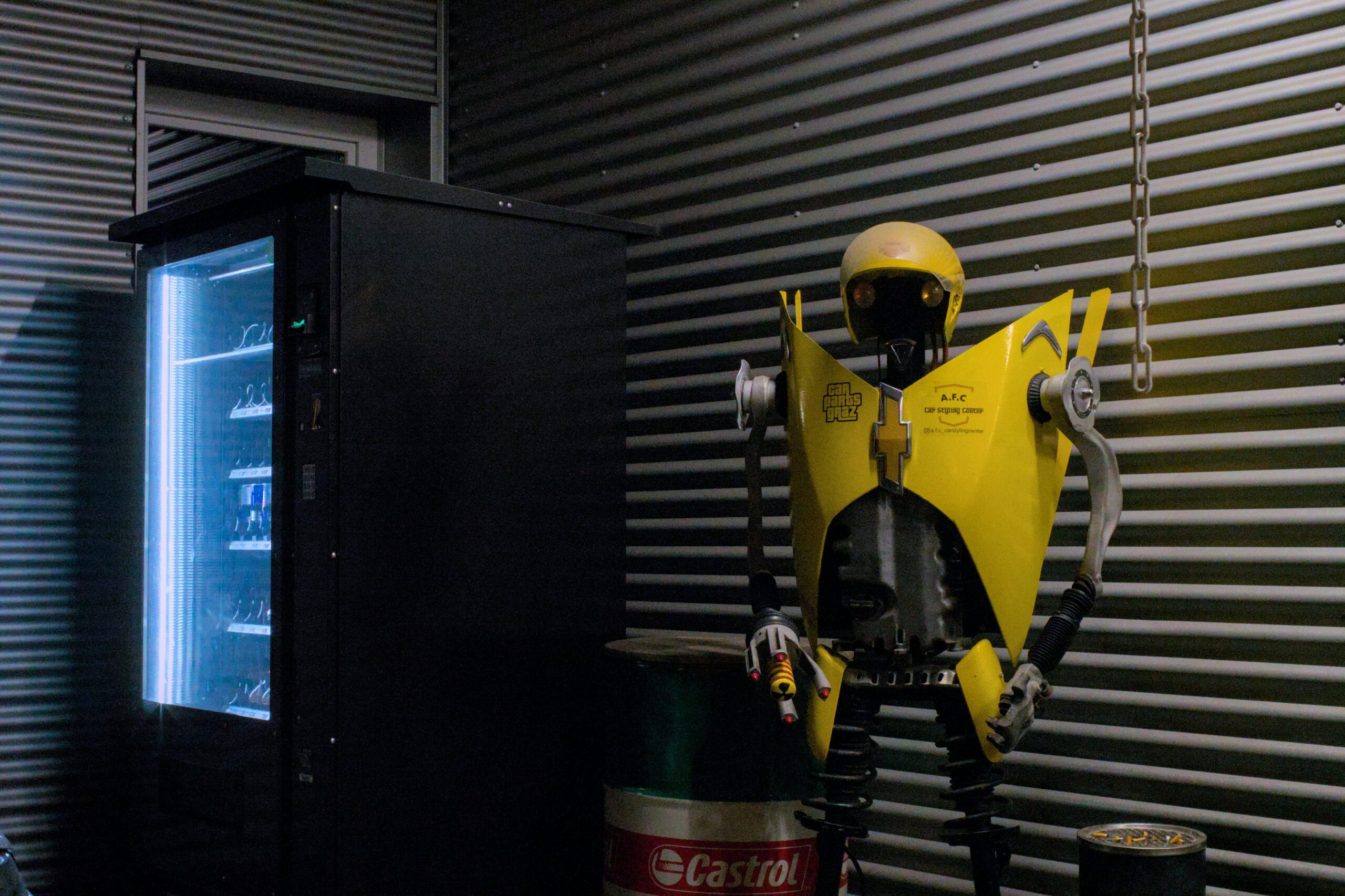 A dimly lit industrial room features a vending machine beside a quirky robot sculpture.