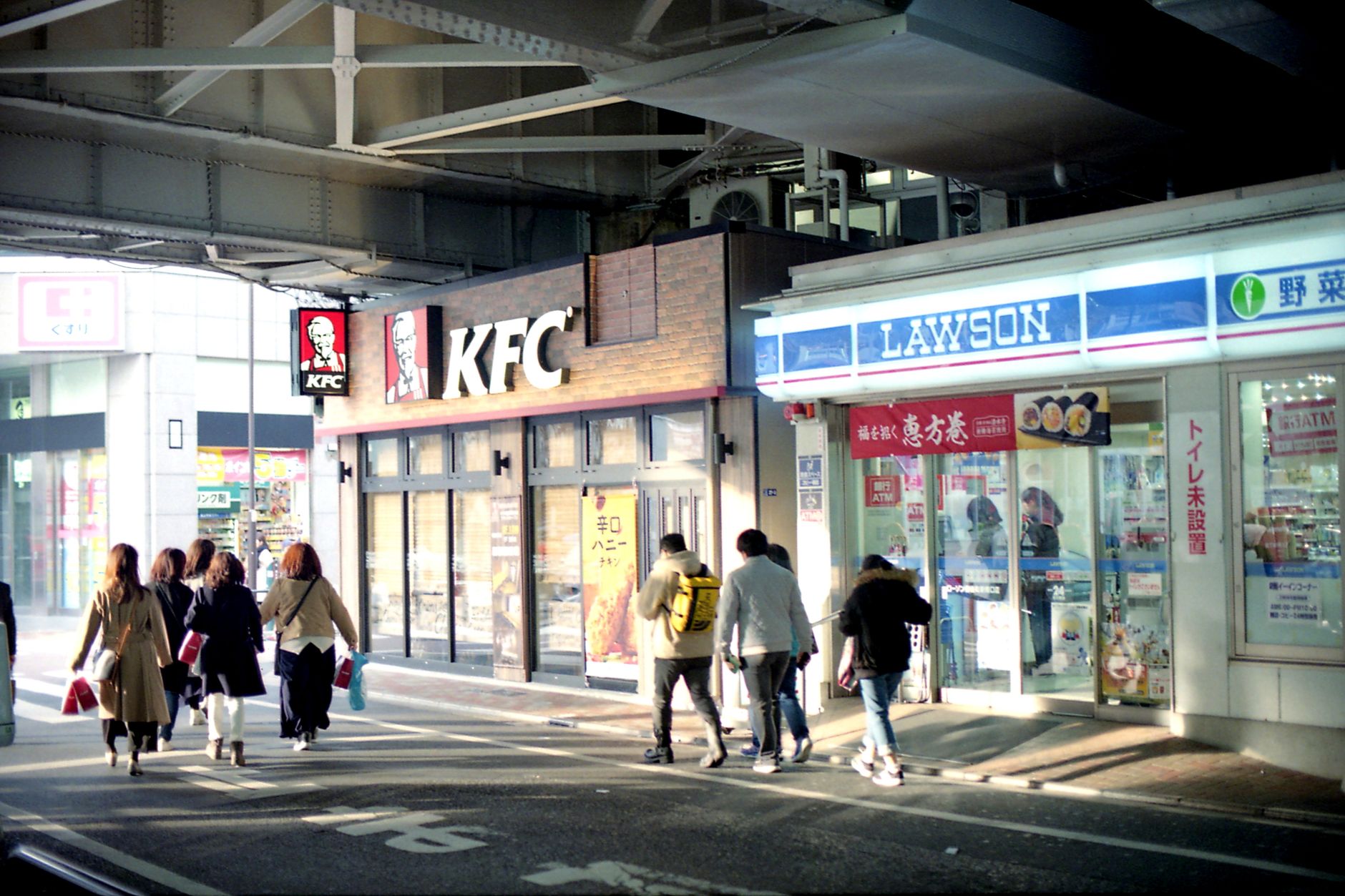 People walking past KFC and Lawson stores under an overpass in Tokyo.