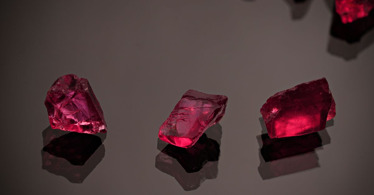 Three red gemstones glitter against a dark reflective background in a still life composition.