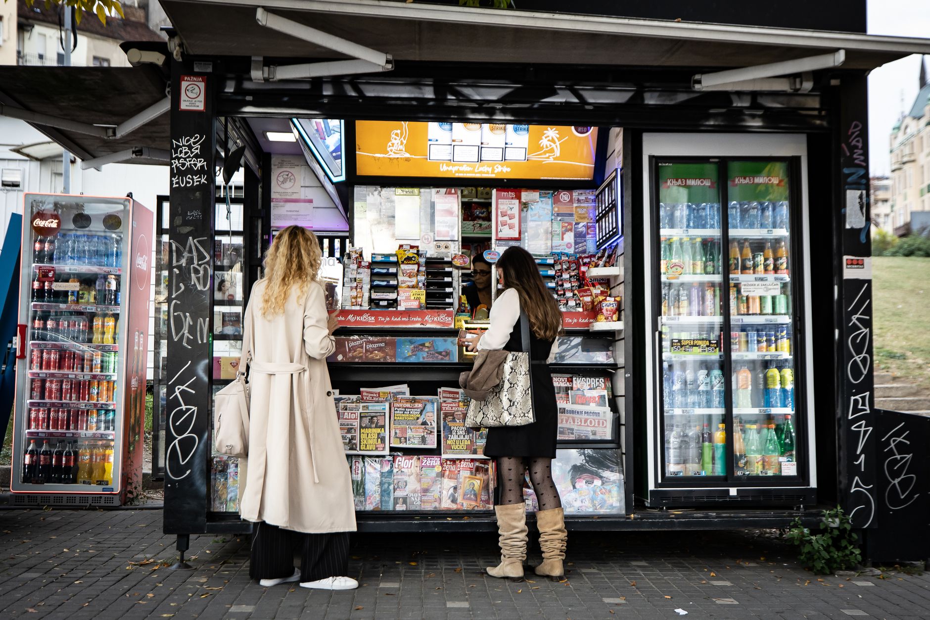 Women browsing at a street kiosk offering magazines and beverages in an urban setting.