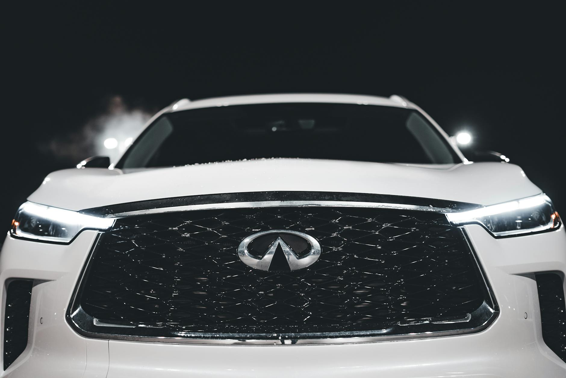 Close-up of a white luxury Infiniti QX60 SUV front grille and headlights at night.