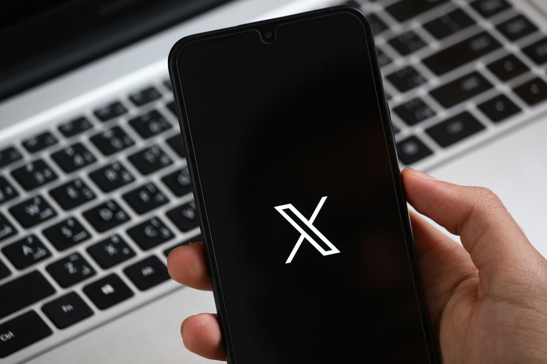 A hand holds a smartphone displaying a modern app interface, with a laptop keyboard in the background.