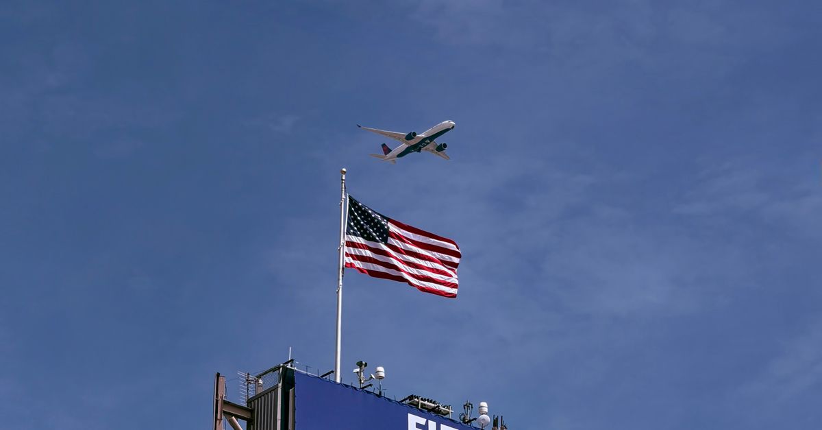 Airplane flying above FIFA World Cup 2026 sign with American flag, clear blue sky.