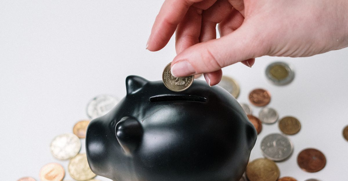Hand inserting coin into black piggy bank among scattered coins, symbolizing savings and financial planning.