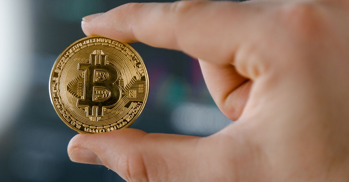 Close-up of a hand holding a Bitcoin against a stock market chart backdrop, symbolizing cryptocurrency investment.