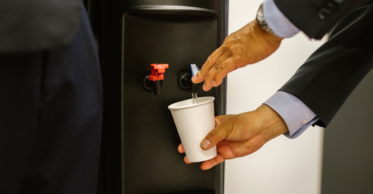 Man in business attire pouring water into a white cup from a dispenser.