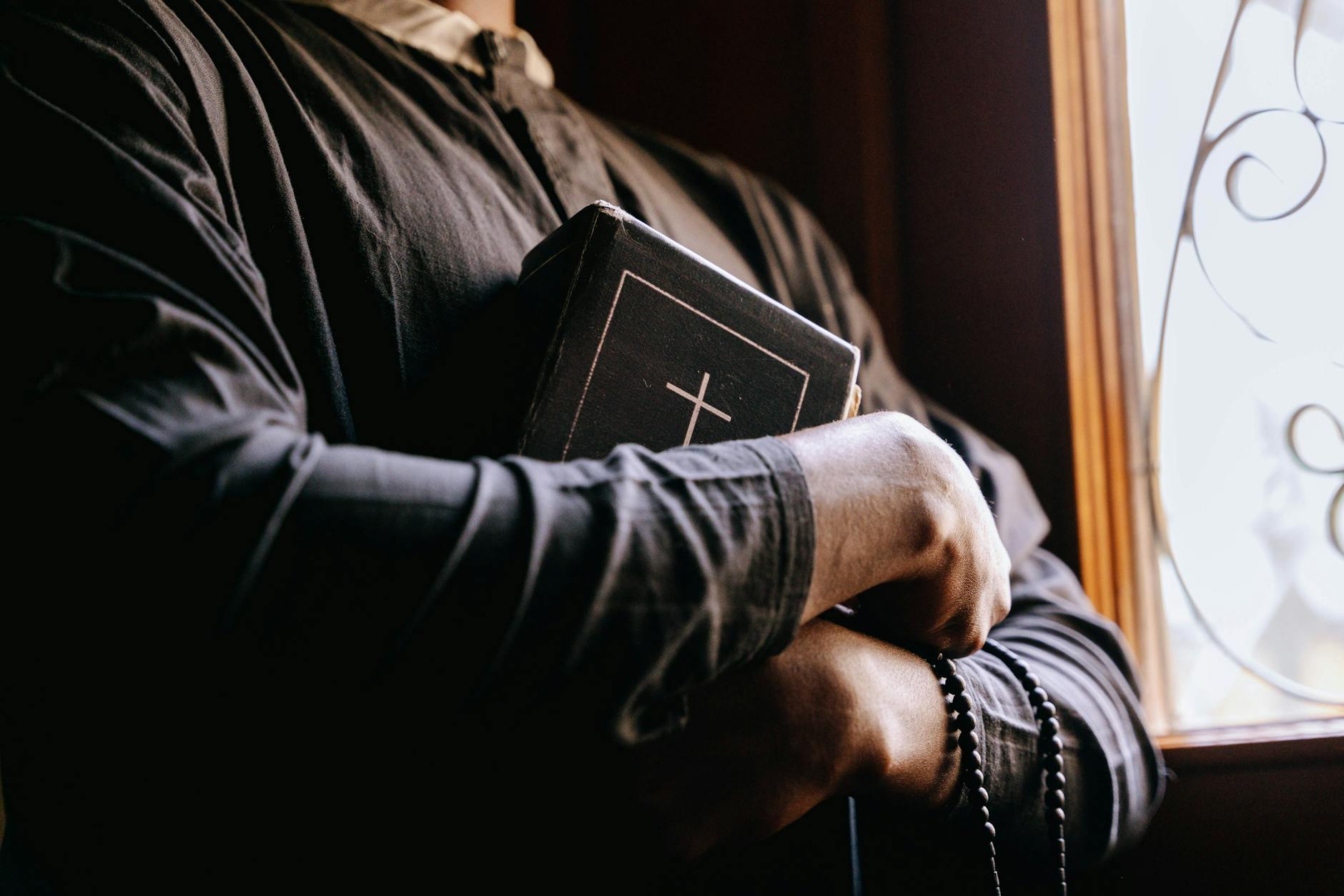 A priest holding a Bible by a sunlit window, symbolizing faith and spirituality.