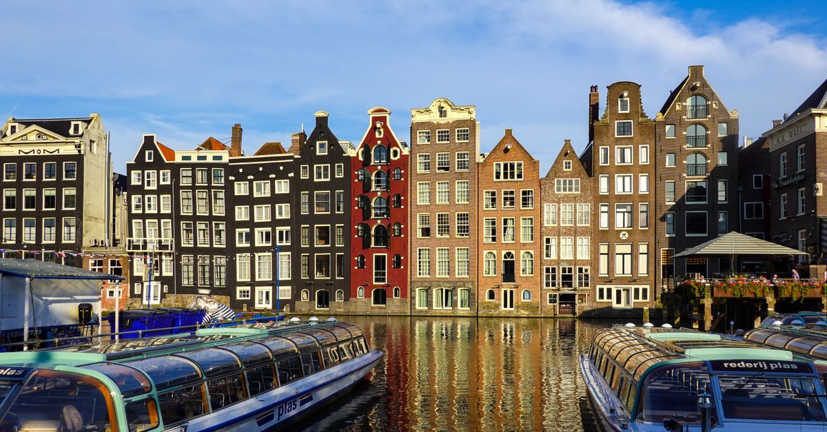 Colorful historic buildings along Amsterdam canals with boats in the foreground, reflecting in calm water.