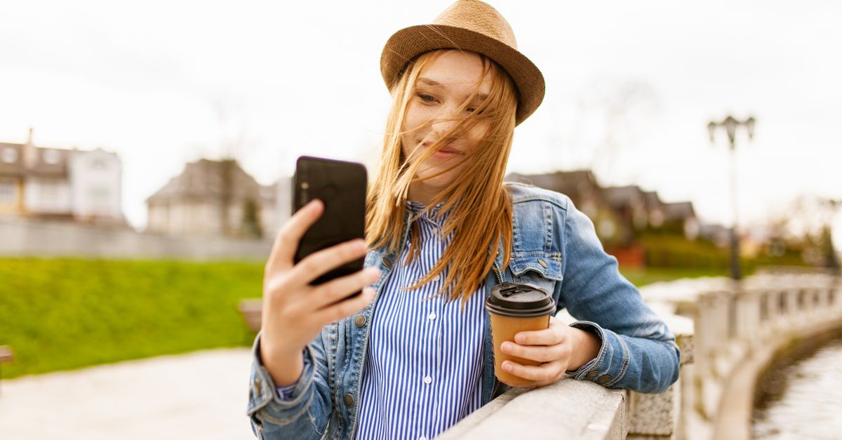 Smiling young woman with coffee outdoors using smartphone by a river.