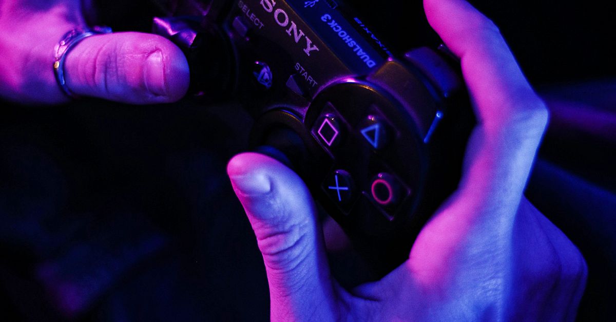 A close-up of hands gripping a Playstation controller, illuminated by purple and blue lighting.