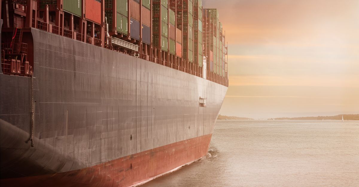 A colossal cargo ship loaded with containers navigates through calm waters against a vibrant sunset sky.