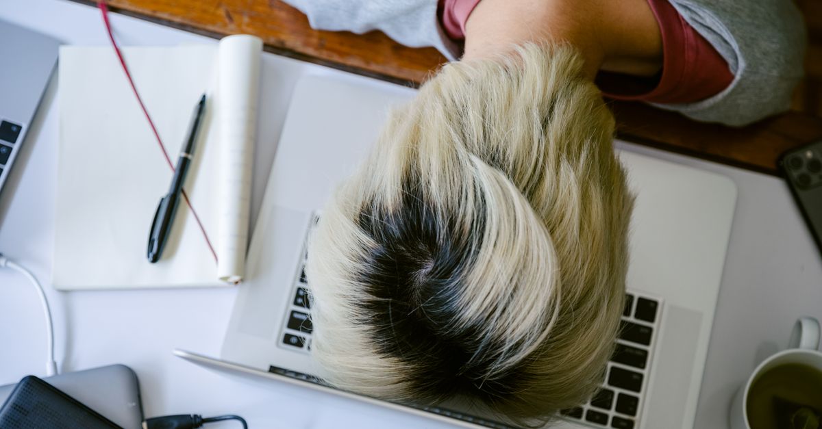 From above of tired fatigued unrecognizable female in casual wear resting head on netbook keyboard near notebook and cup of tea after long working day