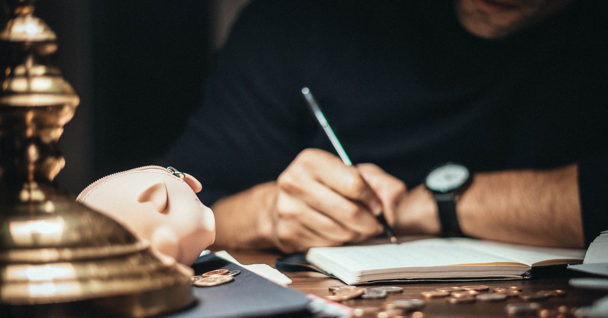 Crop man sitting at desk with vintage lamp and scattered coins and piggybank while writing in notepad
