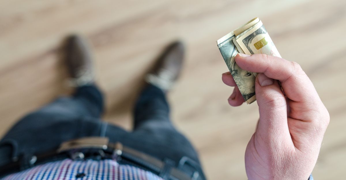 A person wearing denim and a checked shirt holds folded dollar bills in their hand indoors.